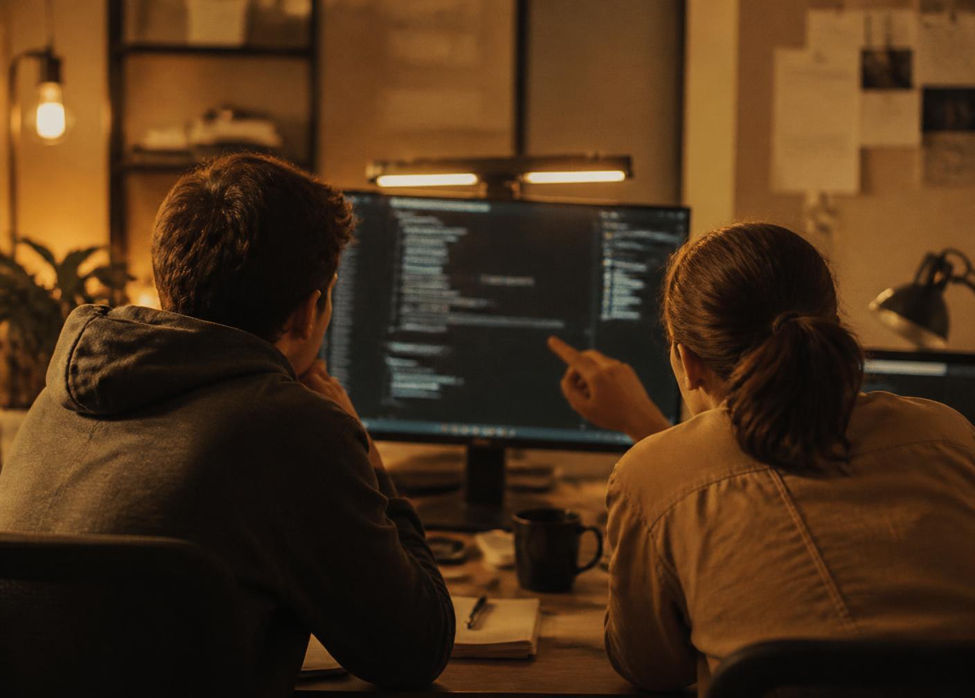 Two engineers discussing code on a monitor together in a warm, low-lit workspace